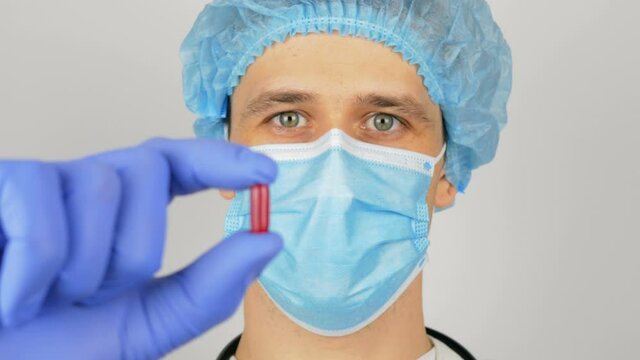 A young handsome doctor in a medical mask holds a red pill in his hands presenting a new drug.