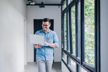 Young cheerful beautiful joyful man manager businessman entrepreneur small business owner employee worker with glasses smiling while working with his laptop walking in office hallway corridor