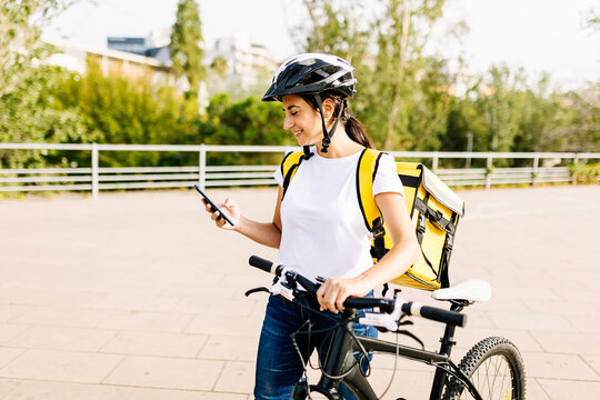 Smiling delivery woman using mobile phone while wheeling bicycle
