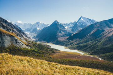 Fototapeta premium Wonderful alpine landscape with mountain lake and mountain river in valley with forest in autumn colors on background of snowy mountains silhouettes under blue sky. Beautiful mountain valley in autumn