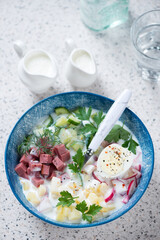 Traditional russian cold soup with kefir or okroshka served in a blue bowl, vertical shot on a beige granite background, elevated view