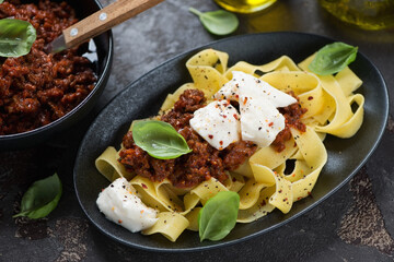 Italian pappardelle pasta served with bolognese sauce, mozzarella and fresh green basil on a black plate, horizontal shot