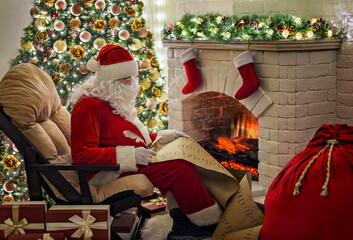Old kind bearded Santa Claus sitting at armchair in his cozy home room near Christmas tree, fireplace, xmas stocking and big gifts sack and writing on kids list preparing for holidays