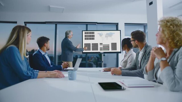Management meeting. People sitting around table and one standing and explaining financial figures on screen. Modern open space office interior.