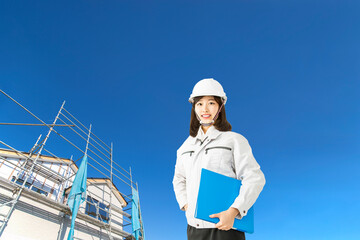 青空と一戸建て工事現場を背景に立つ、作業着を着たカメラ目線の女性