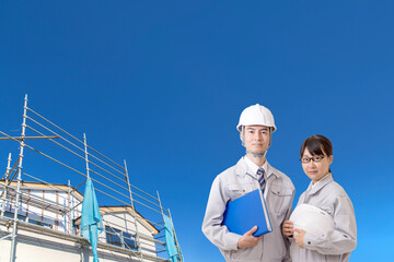 青空と一戸建て工事現場を背景に立つ、作業着を着たカメラ目線の男女技師