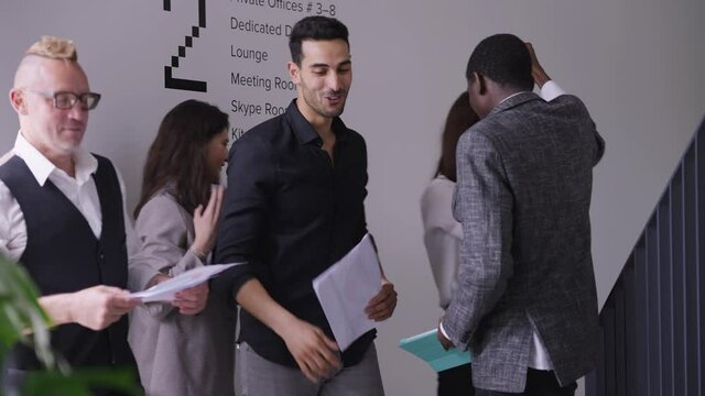 Multiethnic men and women stacking hands smiling and leaving in office. Caucasian Middle Eastern and African American colleagues meeting in hallway walking away to work. Teamwork and confidence