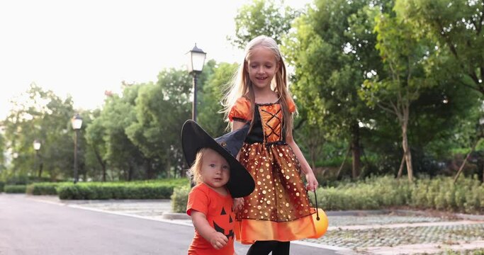 Cute little caucasian kids with blonde hair seven years old in costume of witch with hat and baby one year old celebrating Halloween outdoor on street. Holiday concept. Slow motion.