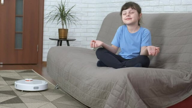 Funny child with a cleaner. A funny young girl relax and do yoga on the sofa while a smart hoover move on the carpet.