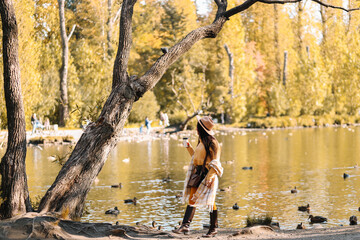 Portrait of a happy cheerful young woman in fashionable casual clothes and a stylish hat walking drinking coffee enjoying solitude in a fall park in nature by the water in autumn, selective focus