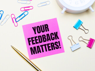 On a light background, a white alarm clock, pink, blue and white paper clips, a golden pen and a pink sticker with the text YOUR FEEDBACK MATTERS