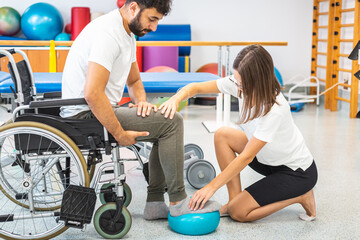 Pphysiotherapist and male patient seated in a wheelchair during rehabilitation treatment - press and release a foot on a small exercise ball