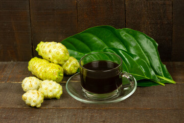 Noni juice in transparent glass and noni fruit with green leaf on wooden background. 