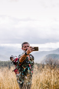 man with dog in backpack taking photos on the mountain with clou