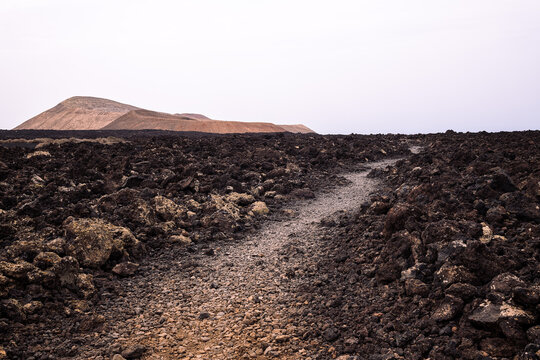 Dry pathway between black lava against volcanoes under light sky