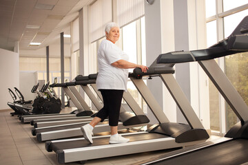 Fototapeta na wymiar elderly retired woman with gray hair is engaged sports on simulators in gym. Healthy lifestyle, senior concept, indoor. Rehabilitation, treatment after injuries, improvement. racecourse