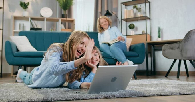 Lovely positive happy blond mother reading book on the couch and watching at her smiling friendly eldest and younger daughters which lying on the floor and applying laptop