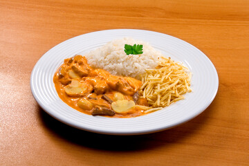 Strogonoff de carne com arroz e batata palha e champignon no fundo de madeira clara.