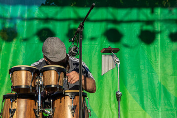 Man with flat cap playing wooden Caribbean bongo drums with mircophone and chimes and green curtain behind