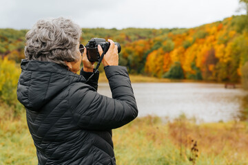 Fototapeta premium Seniors and technology, retirement hobby concept. Back view senior woman takes photo on digital camera, elderly woman tourist takes pictures of autumn beautiful park and lake, copy space