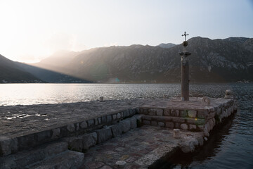 A high stone pillar beacon with a cross and a ball against the backdrop of the Kotor bay, mountains and the last rays of the setting sun. Sea latarnia. The pier. Piled up island. Our Lady of the Rocks