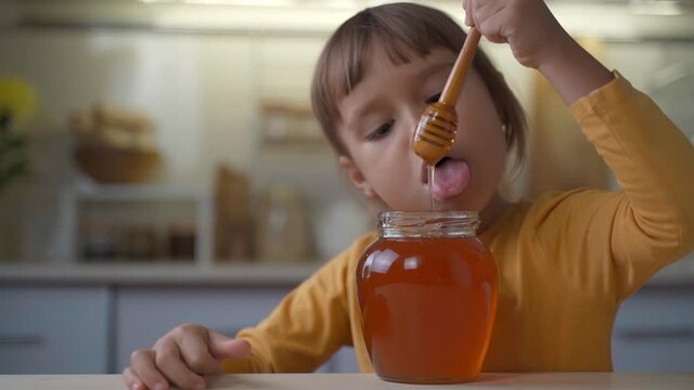 ittle child girl eat honey as it drops on her tongue licking it out from the wooden spoon. Kid eats honey from a jar at home in the kitchen. Healthy organic sweets for kids