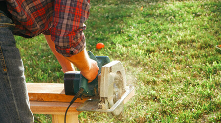 A male carpenter works with a circular saw on the lawn. Close-up view of a working tool. Contrast...