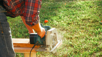 A male carpenter works with a circular saw on the lawn. Close-up view of a working tool. Contrast sunlight with glare scattered in flying sawdust.