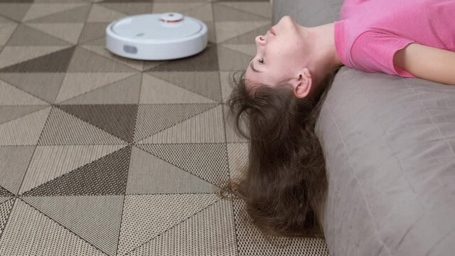 Household robot cleans the house. A cute girl pulled her long hair down to the floor, a robot vacuum cleaner next to her collects dust on the floor.