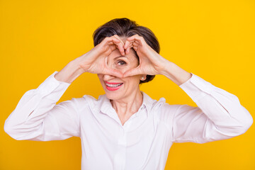 Photo of cute adorable age woman dressed white shirt showing fingers heart cover eye smiling isolated yellow color background