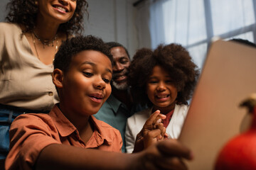 african american boy gesturing during video call on blurred laptop near happy family