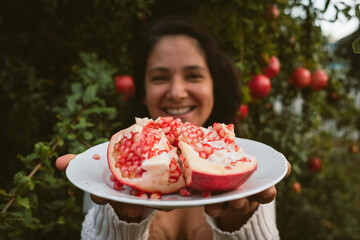 smiling woman hands holding dish with fresh pomegranate pieces harvested from tree in the background with red seeds. concept of eating autumn fruit for healthy diet and lifestyle
