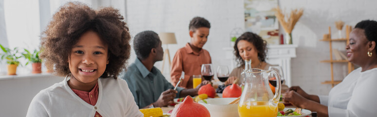 Smiling african american girl standing near blurred family and thanksgiving dinner, banner