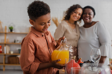 Smiling african american boy pouring orange juice near blurred mother and granny during thanksgiving dinner