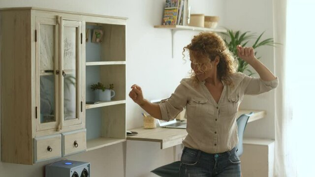 Happy adult woman sing and dance at home in the living room with light and laptop computers in background - happiness and people in smart working workplace - modern people online job creator content