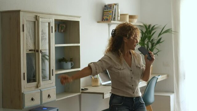 Overjoyed female caucasian people singing and dancing at home in living room with modern workplace in background for smart working activity business lifestyle - excited woman sing with phone