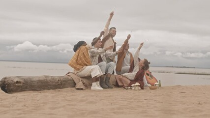 Wide shot of young diverse friends sitting on sandy beach on nasty day, holding sparkling Bengal lights, smiling