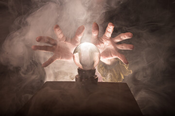 A fortune teller's hands conjure up a crystal ball. Smoke can be seen against a dark background.