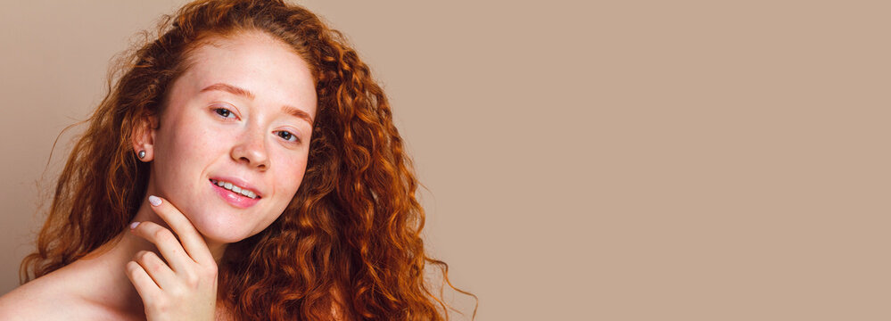 close-up portrait of a beautiful smiling girl with long curly red hair looks on a beige background. No makeup, no retouching, copy space