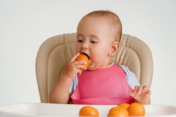 A baby in a pink bib is sitting on a chair trying to eat a tangerine in the peel