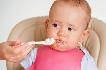A baby in a pink bib contorts his face and does not want to eat porridge with a spoon