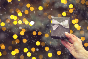 Female hand holding a small gift box at the background of New Year tree during the snowfall