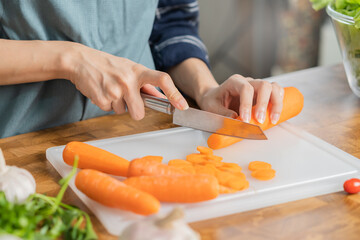 Asian young woman, girl or housewife using knife, cutting carrots on  board, on wooden table in kitchen home, preparing ingredient, recipe fresh vegetables for cooking meal. Healthy food people.