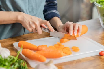 Fotobehang Asian young woman, girl or housewife using knife, cutting carrots on  board, on wooden table in kitchen home, preparing ingredient, recipe fresh vegetables for cooking meal. Healthy food people. © KMPZZZ