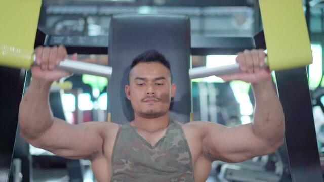 Front View And Close-up Of Young Asian Muscular Fitness Man In A Tank Top Sitting Doing A Chest Workout On An Incline Chest Press Machine By Pulling The Weights Down With Both Hands At A Blurred Gym.