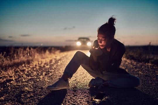 Young woman in jeans and leather jacket sitting in the middle of a road in the countryside with a car lights as background