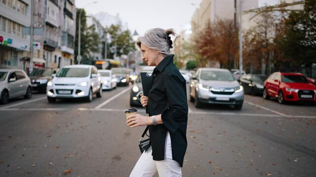 Active adult woman crosses the street at an intersection opposite to the cars standing in a row. Business woman with a folder and coffee in her hands. Slow motion