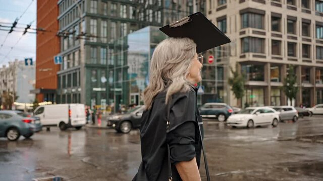 Slow motion of beautiful urban mature woman running down the street in the rain without an umbrella hiding behind a folder with documents. Rain in the city, active aged woman. Slow motion