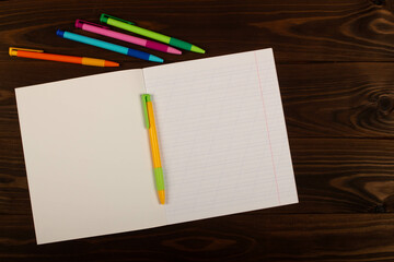 Fototapeta na wymiar School notebook with oblique lines, multicolored pens on the table brown wooden background. Blank sheet of paper in notebook on the desk. Back to school. School supplies. Top view. Copy space for text