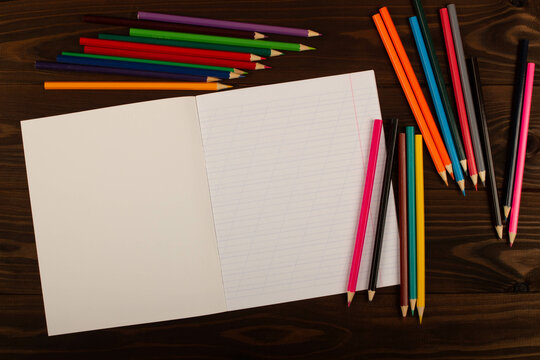 School notebook with oblique lines, colored pencils on the table wooden background. Blank sheet of paper in notebook on the desk. Back to school. School supplies. Top view. Copy space for text